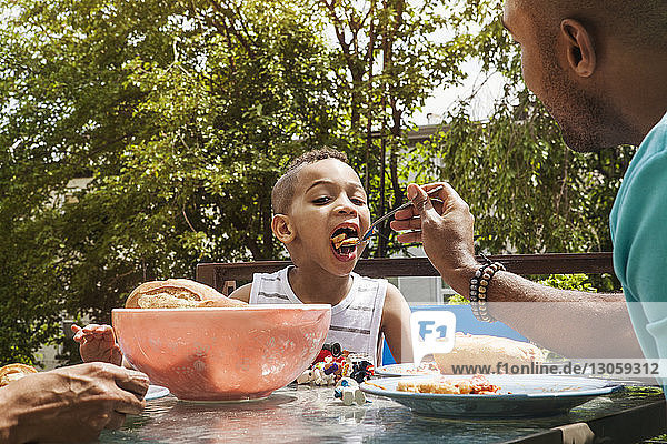 Man feeding son while sitting at table in yard