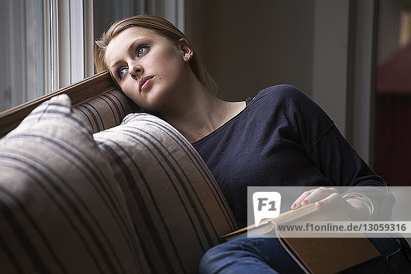 Nachdenkliches Mädchen mit einem Buch in der Hand  das wegschaut  während es sich zu Hause auf dem Sofa entspannt