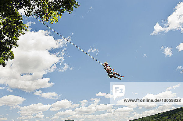 Low angle view of man swinging on rope against sky on sunny day