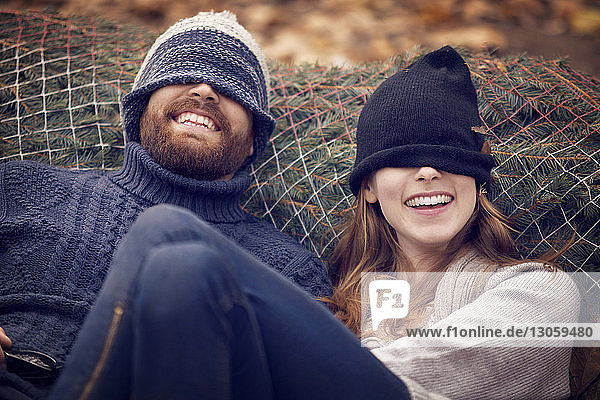 Playful couple wearing knit hat while leaning on Christmas tree in net