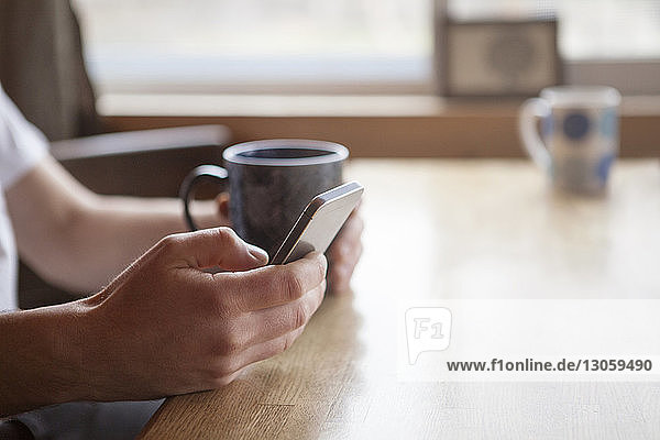 Ausgeschnittenes Bild eines Mannes  der ein Mobiltelefon benutzt  während er im Café eine Kaffeetasse hält