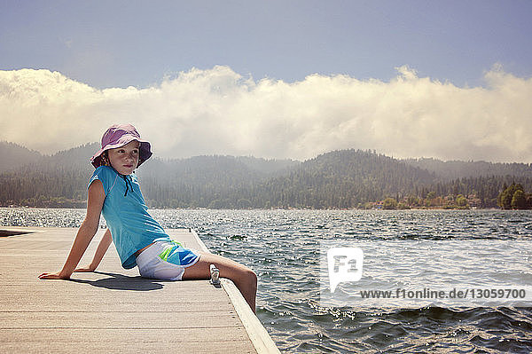 Girl looking away while sitting on pier at lake