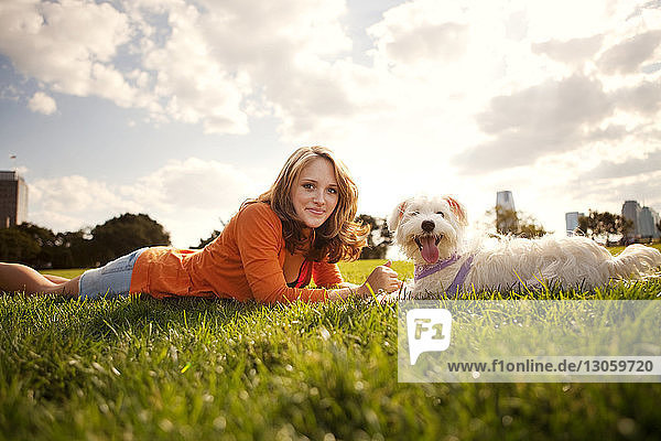 Portrait of young woman lying with dog on grassy field against sky