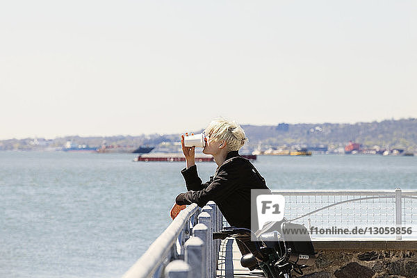 Frau trinkt Kaffee  während sie sich am Beobachtungspunkt am Fluss an ein Geländer lehnt