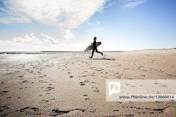 Side view of man with surfboard running on sand at beach against cloudy sky