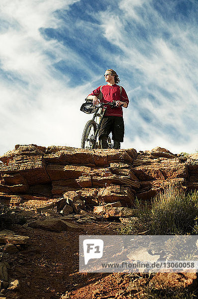 Low angle view of man with bicycle looking away while standing on cliff against sky
