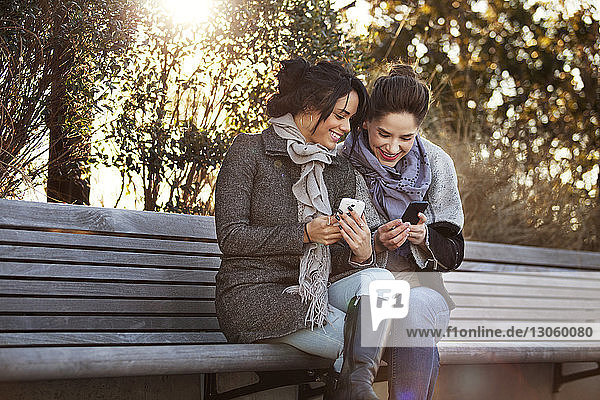 Lächelnde Frauen  die Smartphones benutzen  während sie auf einer Bank sitzen