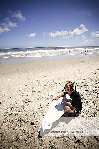 Hochwinkelaufnahme eines Mannes mit Surfbrett  der am Strand im Sand kauert