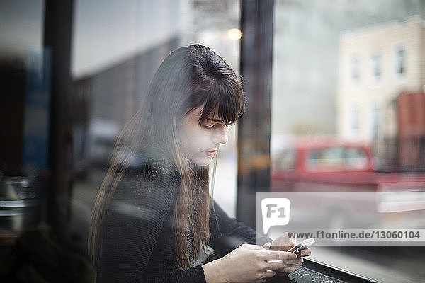 Nahaufnahme einer jungen Frau  die ein Smartphone benutzt  durch ein Fenster gesehen
