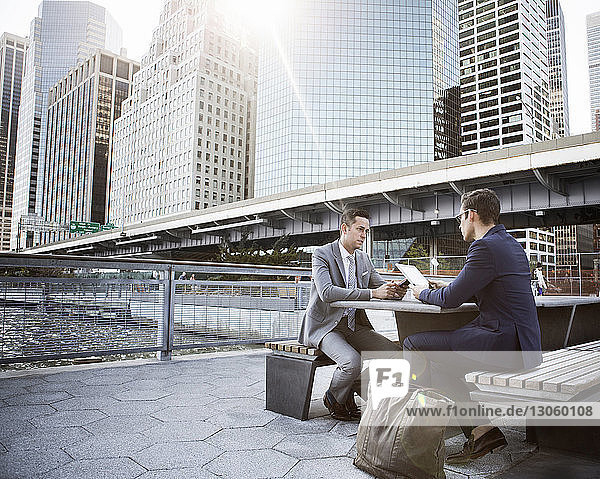 Businessmen using tablet computer and mobile phone while sitting at bench in city