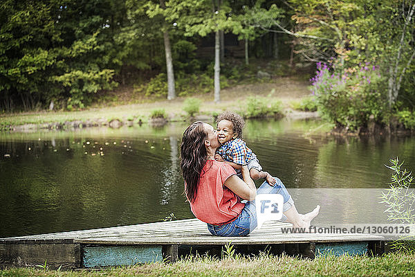 Mother kissing son while sitting on jetty at lake