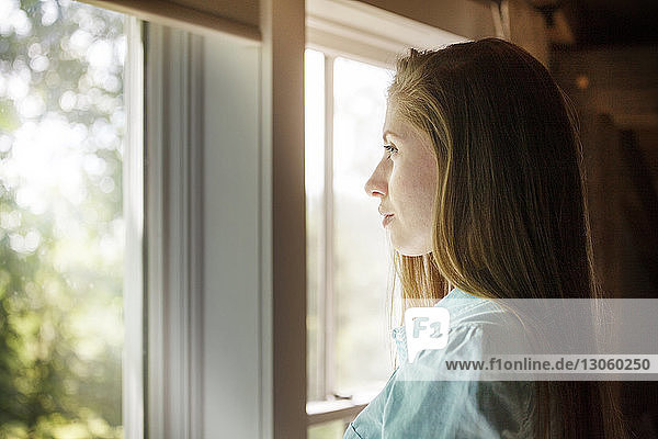 Seitenansicht einer Frau  die zu Hause durch ein Fenster schaut
