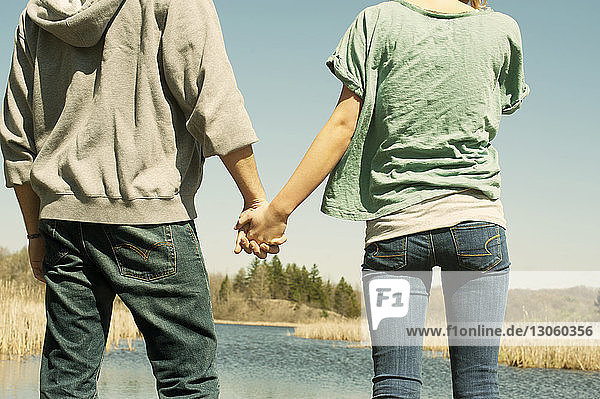 Rear view midsection of couple holding hands while standing at riverbank against clear sky