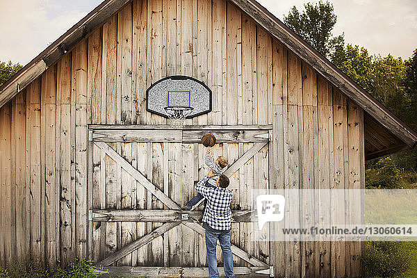 Rückansicht eines Vaters  der einen Sohn trägt  während er vor der Scheune Basketball spielt