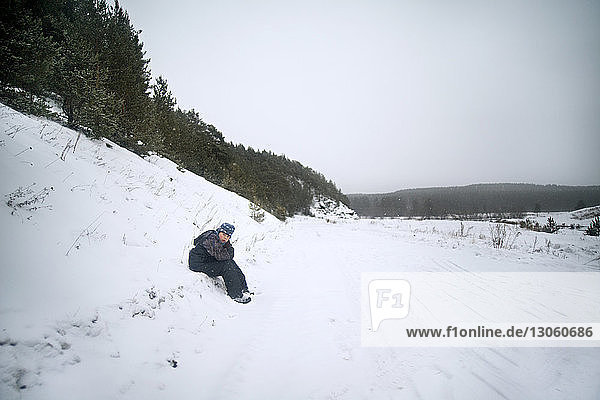 Teenage boy sitting on snow covered field against sky