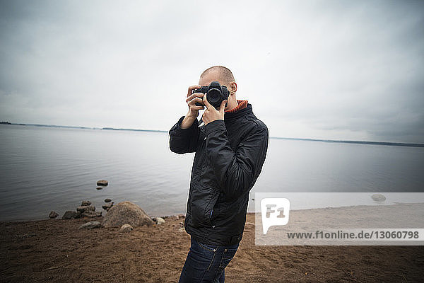 Man photographing with camera while standing on lakeshore against cloudy sky