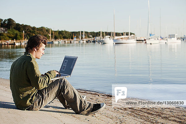 Mann benutzt Laptop-Computer  während er am Strand vor klarem Himmel sitzt