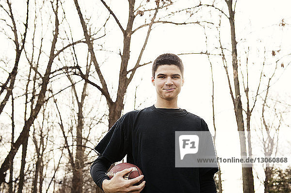 Portrait of smiling boy holding American football against bare trees