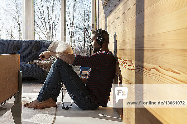 Seitenansicht eines Mannes  der Musik über Kopfhörer hört  während er zu Hause an der Wand sitzt