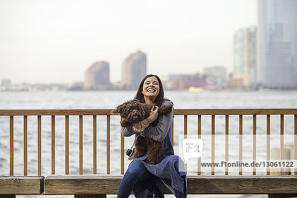 Verspielte Frau mit Hund auf Bank sitzend gegen Hudson River