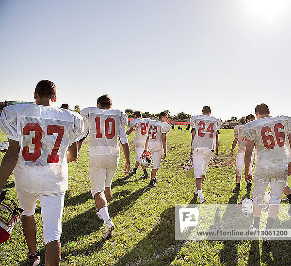 Rear view of American football players walking on field against clear sky