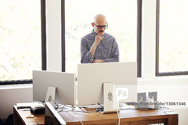 Nachdenklicher Geschäftsmann schaut auf Computer  während er im Büro am Fenster steht