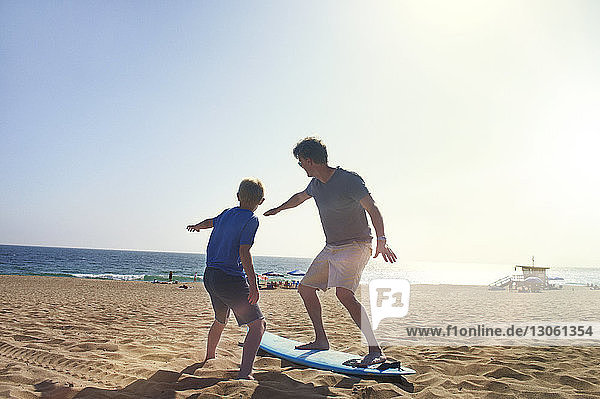 Father and son with surfboard on sand at beach against clear sky