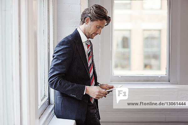 Seitenansicht eines Geschäftsmannes  der ein Tablett benutzt  während er sich im Büro am Fenster lehnt