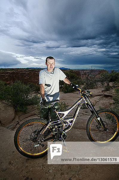 Portrait of man with bicycle standing on rocks against cloudy sky
