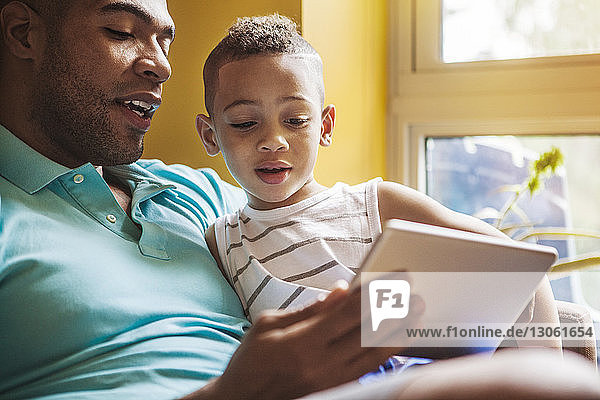 Mann zeigt seinem Sohn einen Tablet-Computer  während er zu Hause auf dem Sofa sitzt