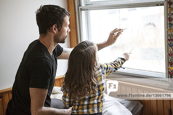 Father and son drawing on window glass while kneeling on bed at home