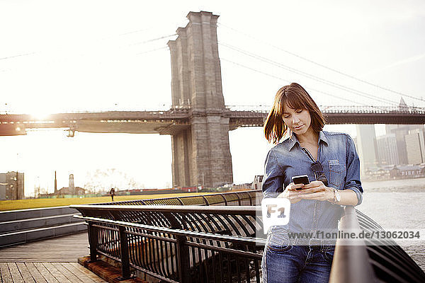 Frau sendet Textnachrichten  während sie am Geländer gegen die Brooklyn Bridge steht