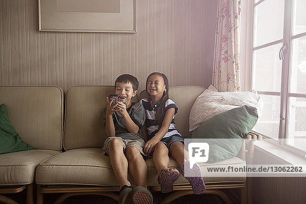 Happy siblings using phone while relaxing on sofa