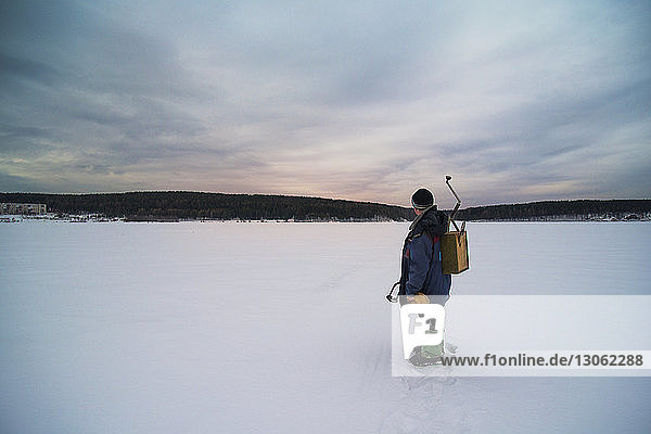 Male holding equipment while standing on frozen lake against cloudy sky