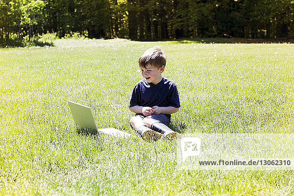 Glücklicher Junge schaut auf Laptop-Computer  während er auf einem Grasfeld im Park sitzt
