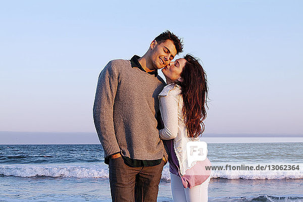 Happy woman kissing man while standing at beach against clear sky