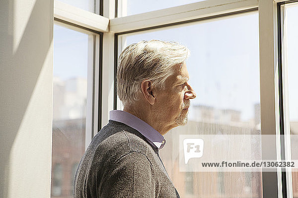 Seitenansicht eines älteren Mannes  der zu Hause durch ein Fenster schaut