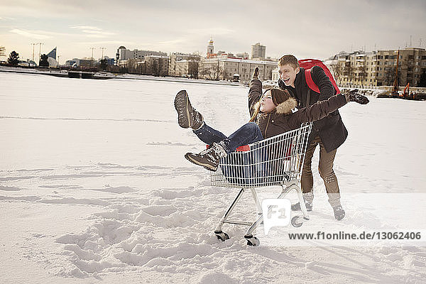 Happy man pushing girlfriend sitting in shopping cart on snow covered field