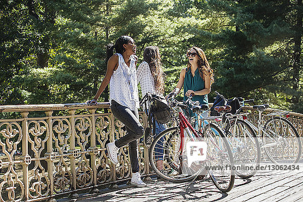 Cheerful friends standing with bicycle by railing on sunny day