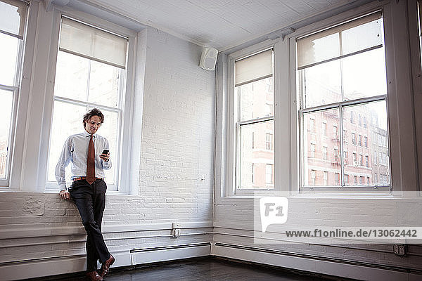 Geschäftsmann benutzt Mobiltelefon  während er sich im Büro ans Fenster lehnt