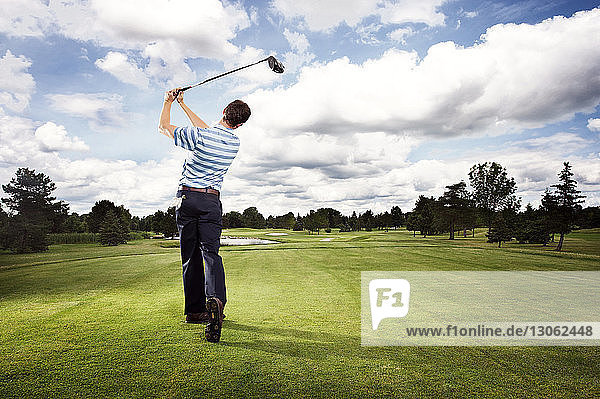 Full length rear view of man swinging golf club on field against cloudy sky