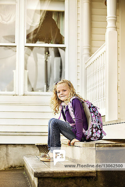Lächelndes Mädchen trägt Rucksack  während es auf Stufen vor dem Haus sitzt