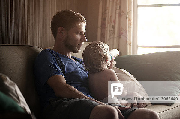 Father sitting with son drinking milk on sofa at home