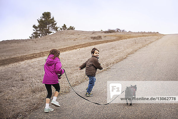 Geschwisterkind mit Hund rennt auf Straße durch Feld gegen klaren Himmel