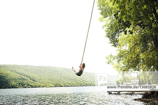Woman swinging on rope over lake against clear sky
