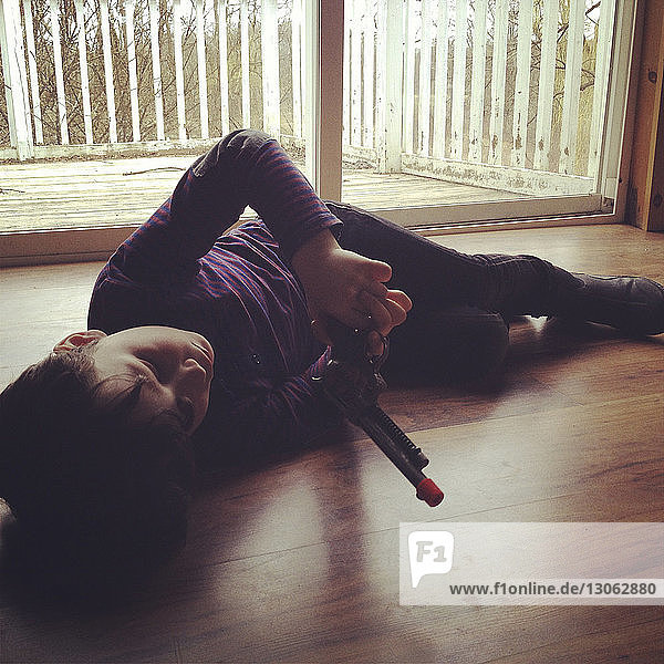 Junge hält Spielzeugpistole in der Hand  während er zu Hause auf Hartholzboden liegt