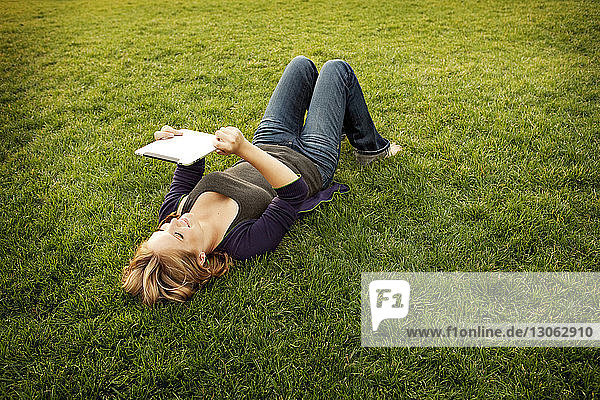 Hochwinkelansicht einer glücklichen Frau  die ein digitales Tablett benutzt  während sie auf einem Grasfeld liegt