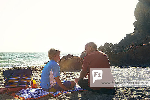 Rückansicht eines älteren Mannes  der mit seinem Enkel spricht  während er an einem sonnigen Tag am Strand sitzt