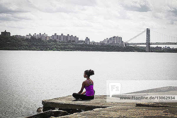 Rear view of woman exercising while sitting on pier at riverbank