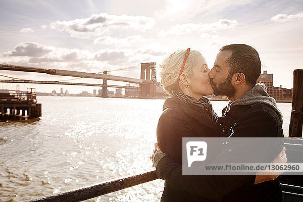 Ein Paar küsst sich  während es an einem sonnigen Tag an der Brooklyn Bridge über den East River steht